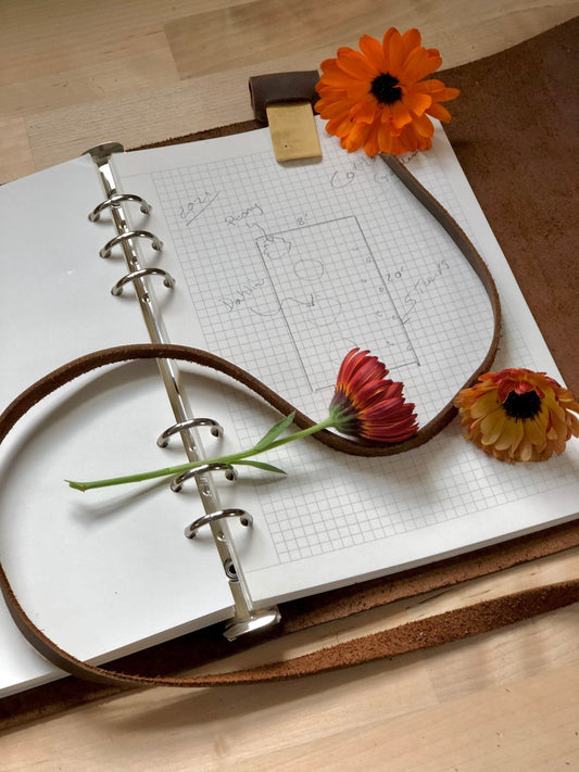 The Celtic Farm’s Leather Garden Journal and Diary, designed for garden thoughts, planning, and design, rests open on a wooden surface—graph pages filled with notes and three orange flowers. A brown strap loops across the cover.