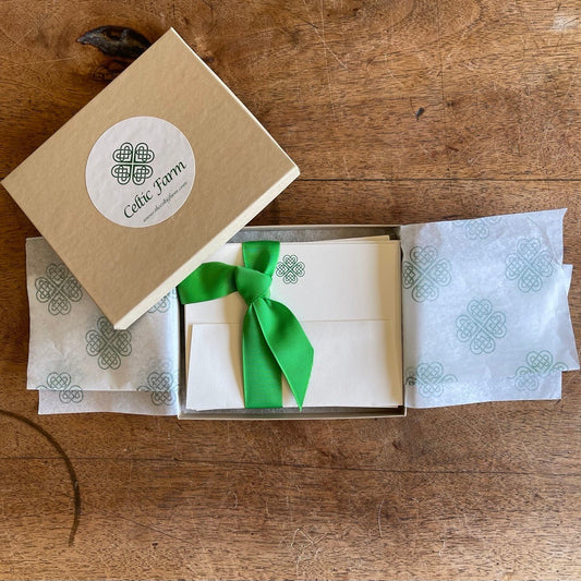 An open box of The Celtic Farm’s Boxed Celtic Knot Stationery reveals 10 white Irish note cards and envelopes, tied with green ribbon, plus tissue paper printed with green Celtic Clover Knots. The lid rests beside the box on a wooden surface.