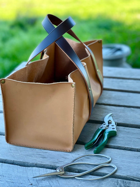 The Celtic Farm Leather Gardeners Tool Bag with dark brown handles sits on a wooden table outdoors, surrounded by green-handled pruning shears and vintage-style scissors—an ideal gardening gift amid lush greenery.