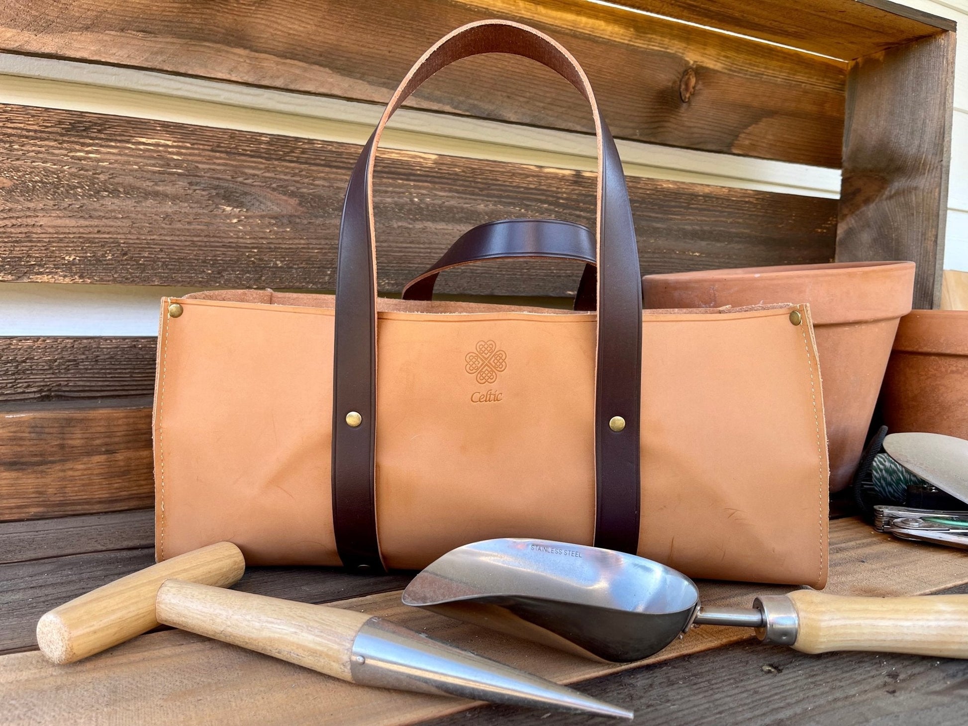 The Celtic Farm Leather Gardeners Tool Bag, in tan leather with dark brown handles, rests on a wooden surface among a trowel, dibber, metal scoop, and terracotta pots—an ideal gift for gardening enthusiasts.
