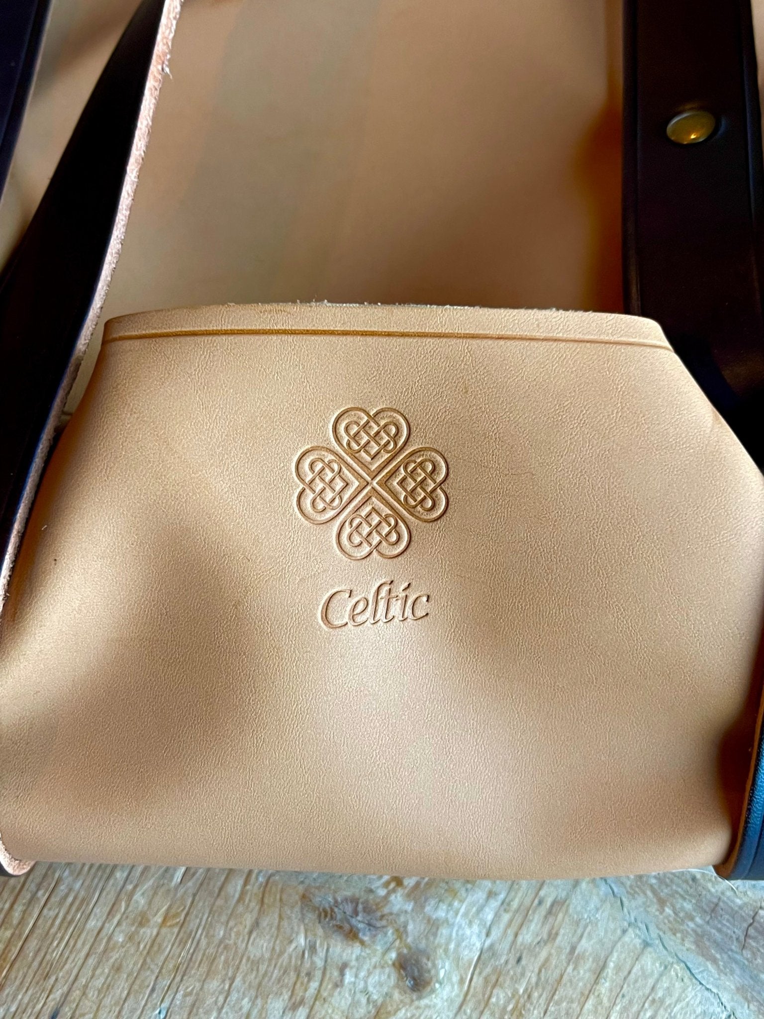 A close-up of The Celtic Farm’s light tan Leather Gardeners Tool Bag with a Celtic knot and “Celtic” embossed, dark leather accents, and a brass rivet—an ideal gardening gift, resting on a wooden surface.