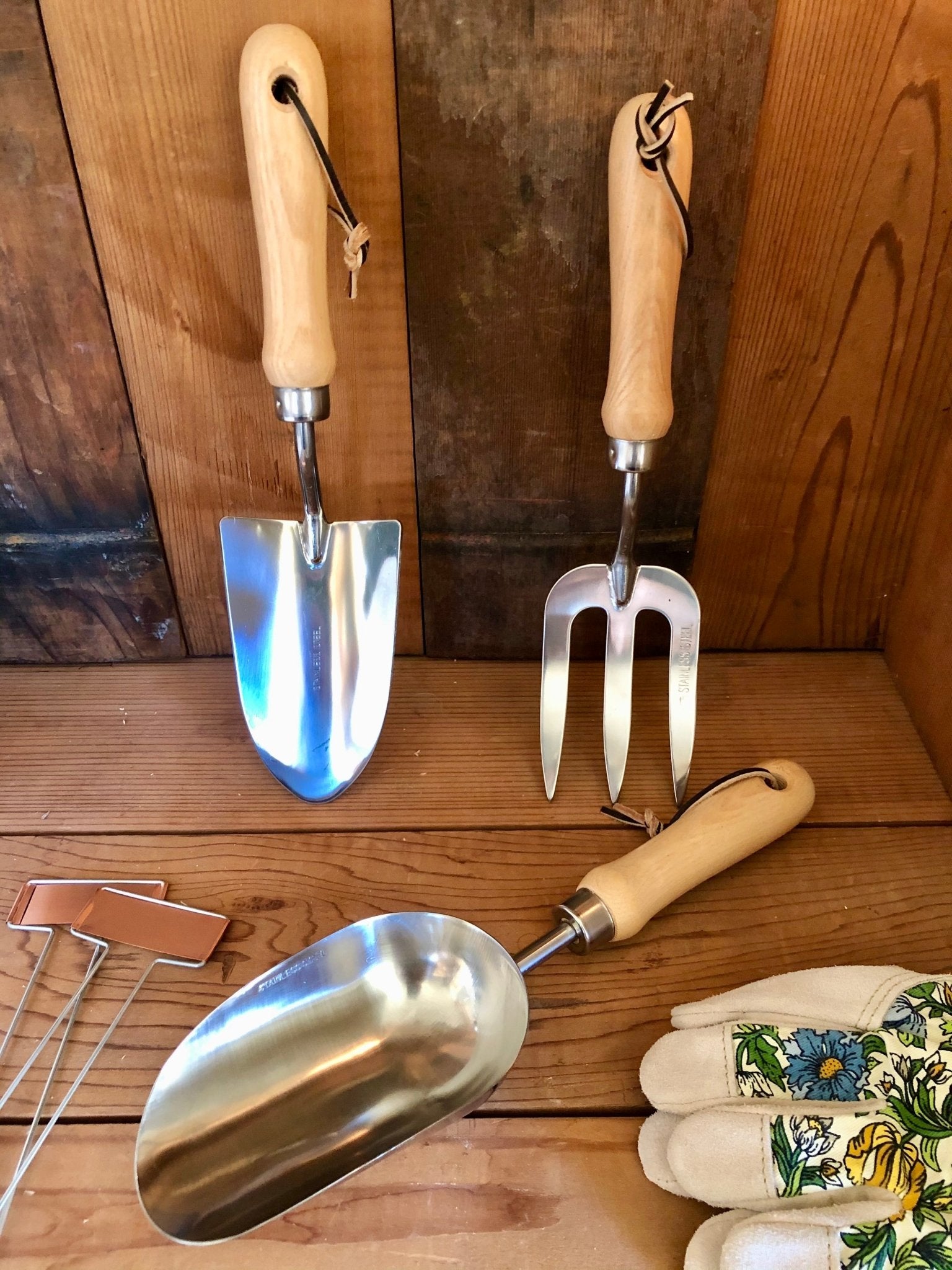 The Celtic Farms Garden Hand Tool Set - Hardwood and Stainless features two trowels and a fork with wooden handles, arranged with labeled plant markers and floral gloves to create the essential gardening kit.