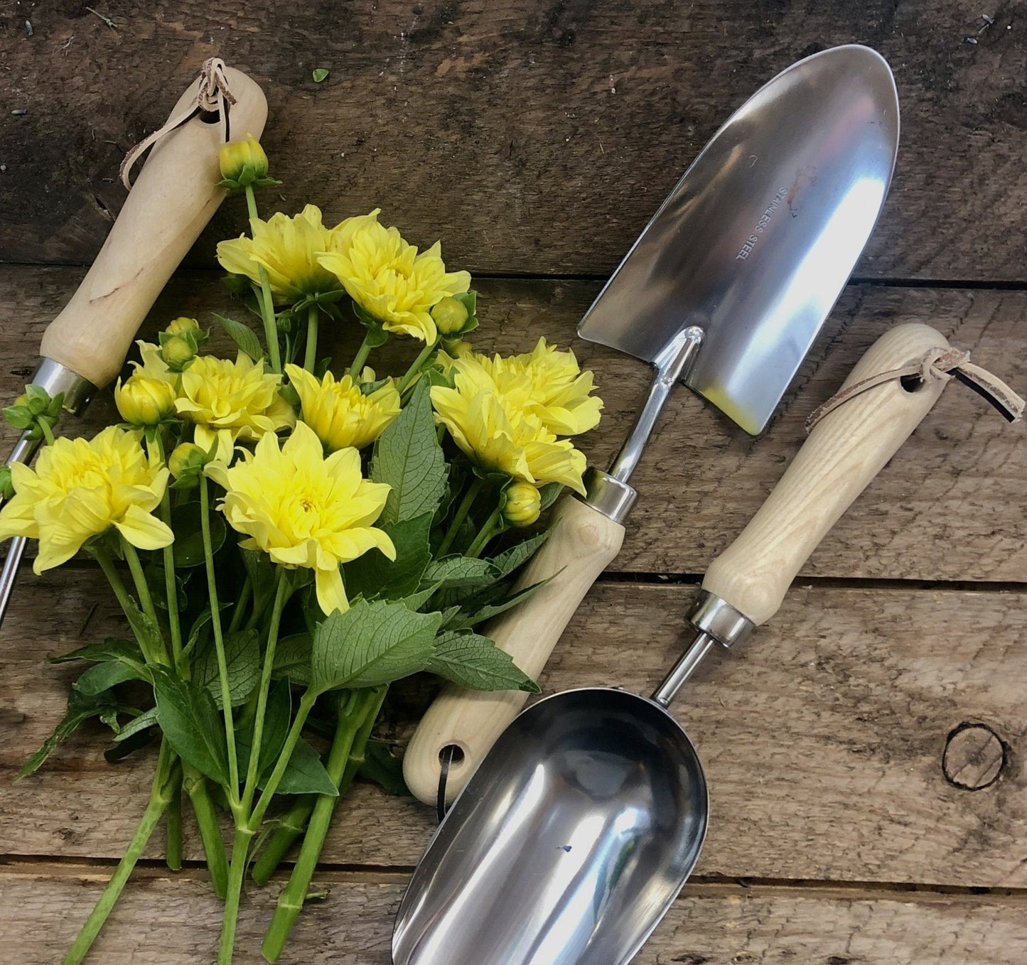 A bunch of yellow flowers rests on wood beside The Celtic Farm’s Garden Hand Tool Set - Hardwood and Stainless, featuring a trowel, transplanter, and hand scoop with wooden handles.