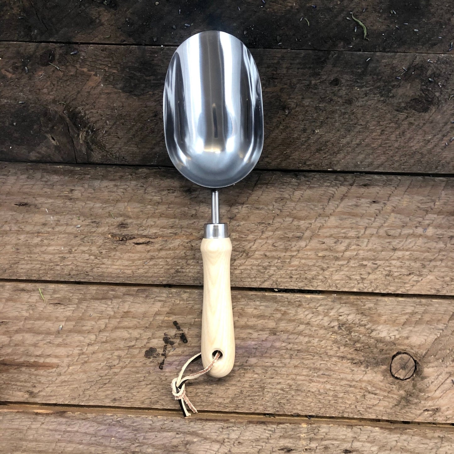 The Celtic Farm Garden Hand Tool Set - Hardwood and Stainless includes a shiny stainless steel scoop with a light wood handle and leather strap, displayed on rustic wood—showcasing the set’s high-quality craftsmanship.