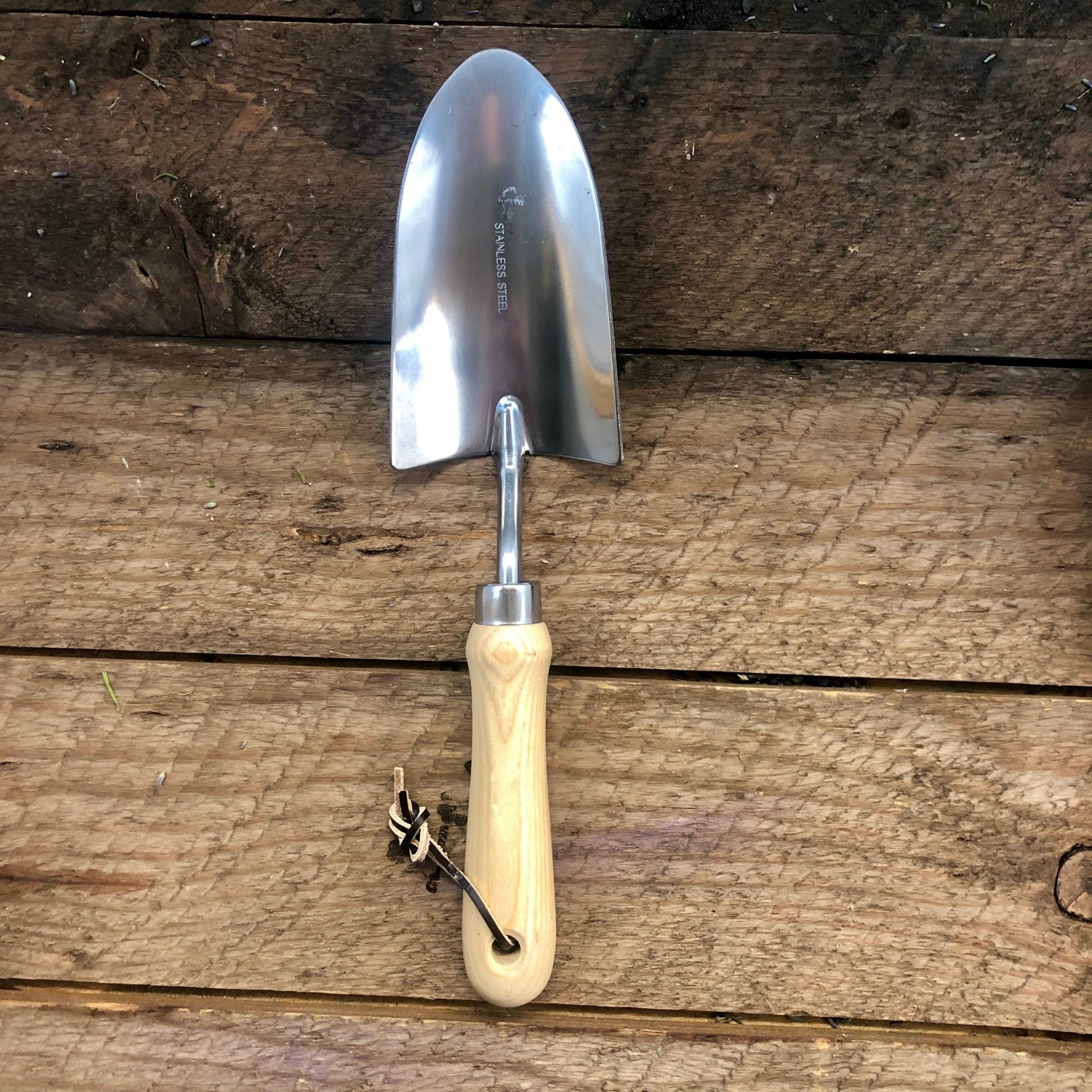 The Celtic Farm’s Garden Hand Tool Set - Hardwood and Stainless features a small garden trowel with a stainless steel blade, light wooden handle, and leather strap, elegantly displayed on a rustic wooden surface.