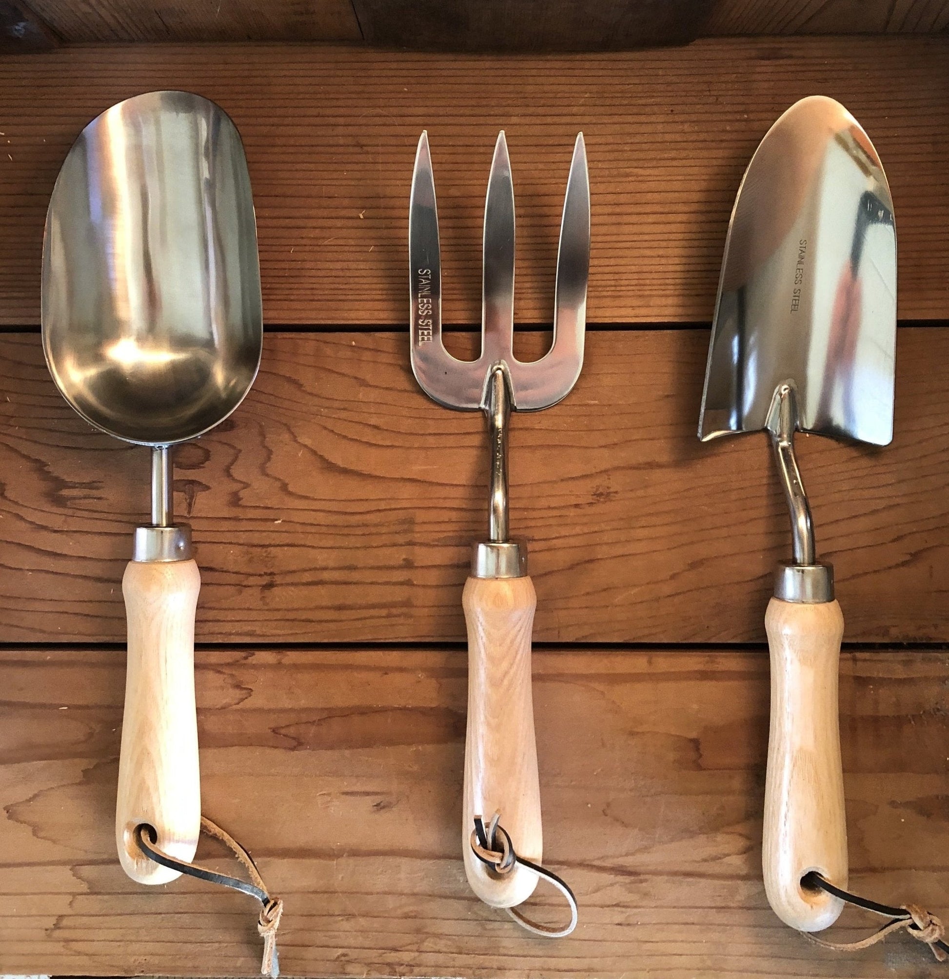 The Celtic Farm Garden Hand Tool Set - Hardwood and Stainless features a scoop, fork, and trowel with stainless steel heads, light wooden handles, and leather loops, neatly hanging side by side on a wooden wall.