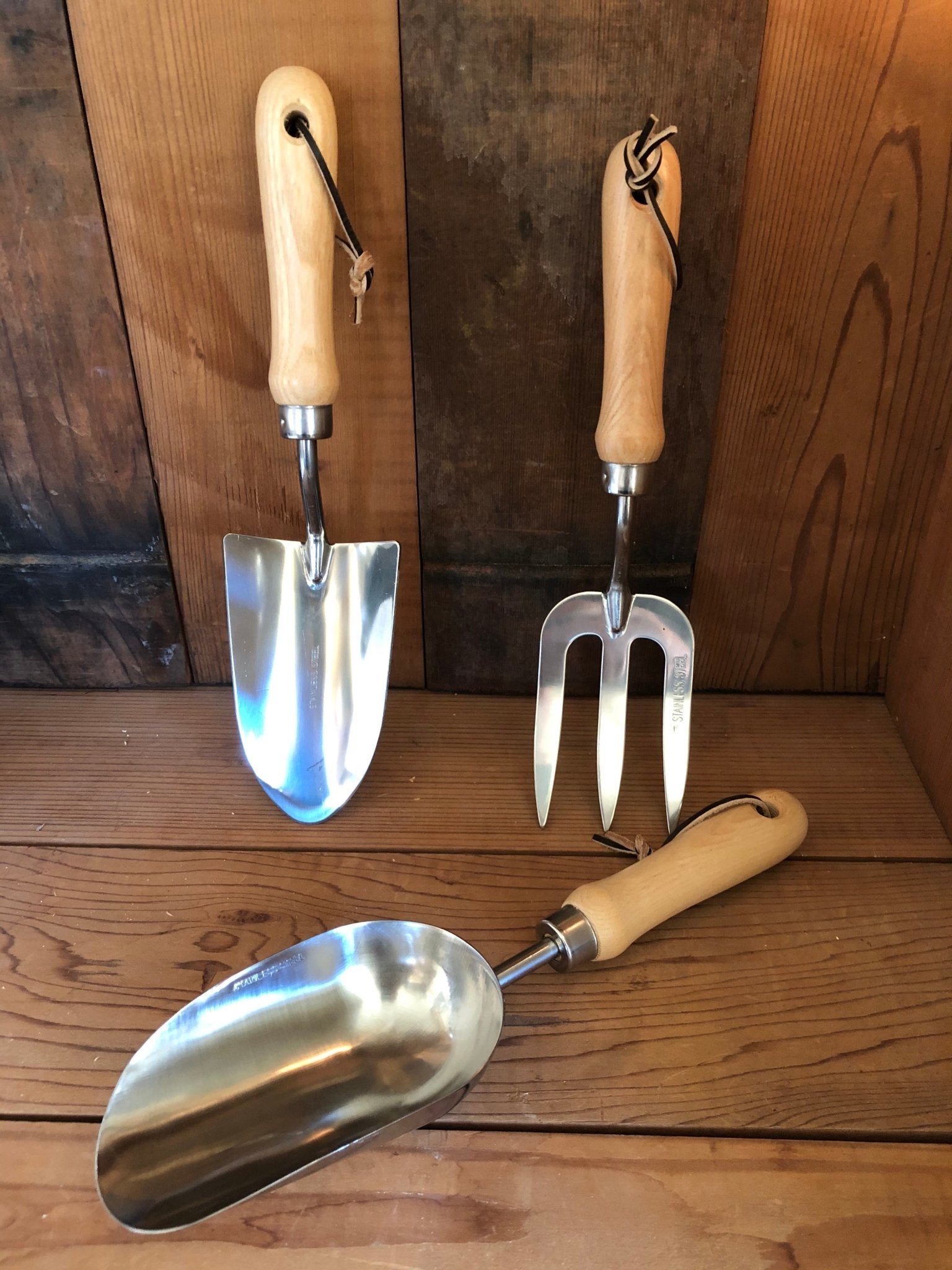 The Celtic Farms Garden Hand Tool Set - Hardwood and Stainless includes a stainless steel trowel, hand fork, and scoop with wooden handles, shown upright and lying on a wood surface against a wood-paneled background.