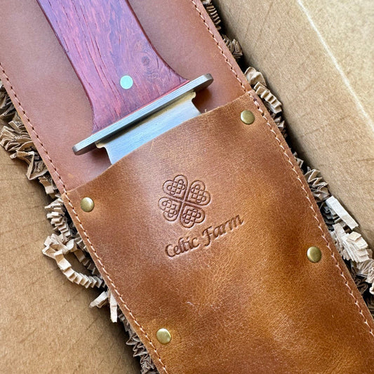 A close-up of the Hori Hori Garden Gift Box by The Celtic Farm shows the garden knife with a wooden handle in a brown leather sheath, embossed with a Celtic knot and The Celtic Farm, resting on shredded paper inside its box.