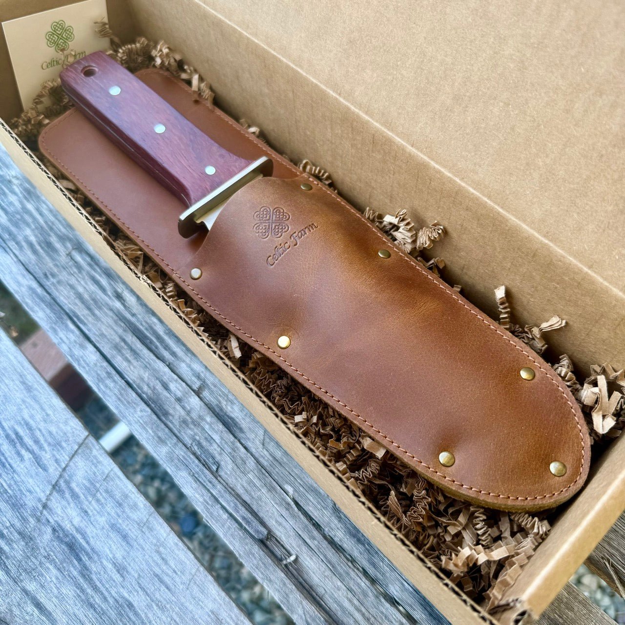 The Celtic Farms Hori Hori Garden Gift Box includes a garden knife with a wooden handle and leather sheath, presented in a cardboard gift box lined with shredded packing material on a wooden surface.