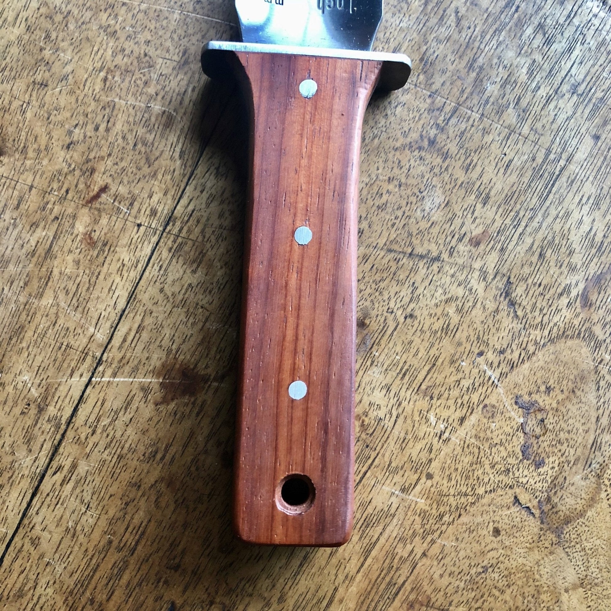 Close-up of The Celtic Farms Hori Hori Garden Gift Box knife handle, featuring three metal rivets and a small hole at the bottom, resting on a scratched wooden surface.
