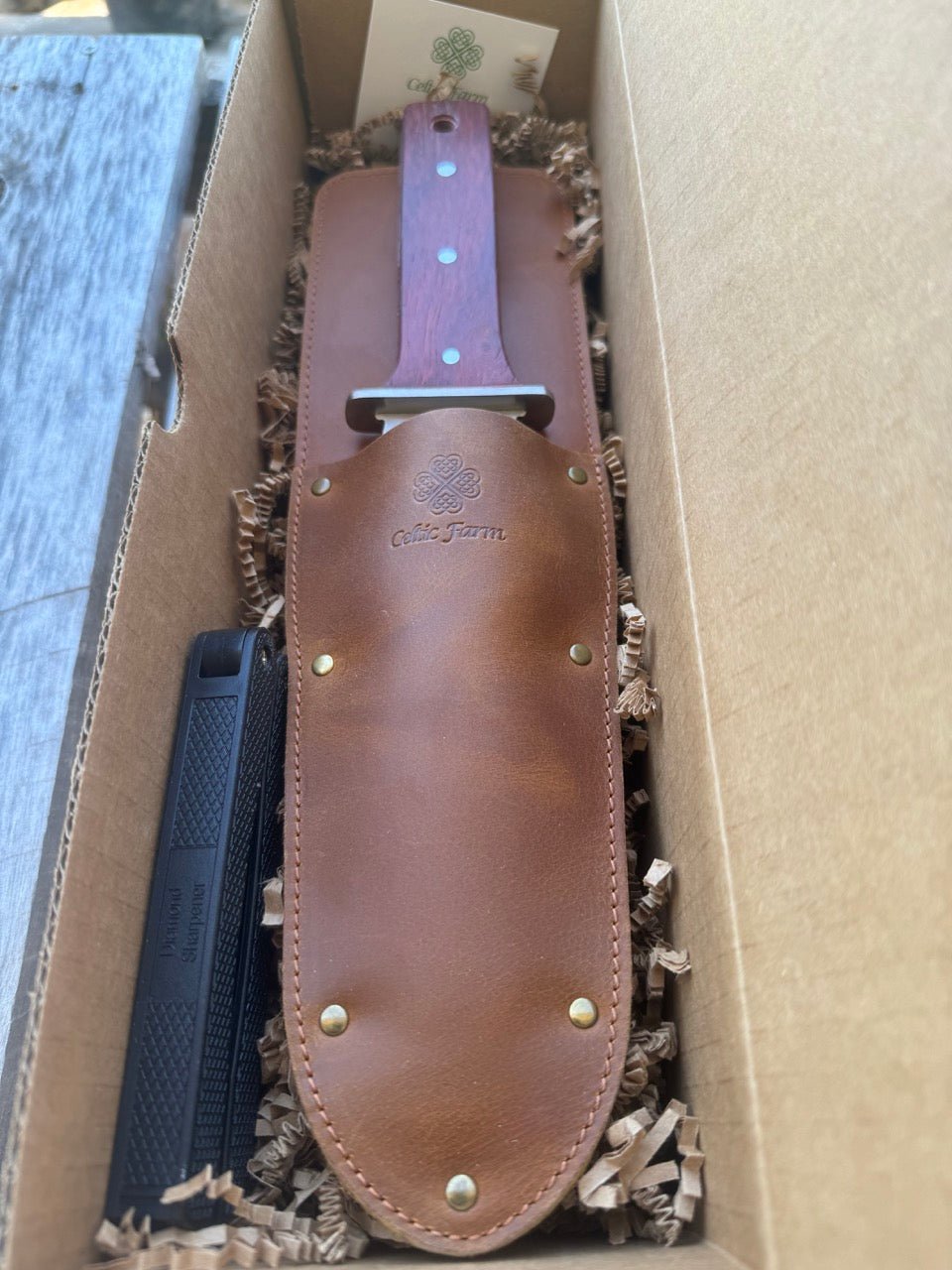 The Celtic Farm Hori Hori Garden Gift Box includes a large garden knife with a wooden handle in a brown leather sheath, packed in a paper-filled box along with a diamond sharpener and a card featuring the green logo.