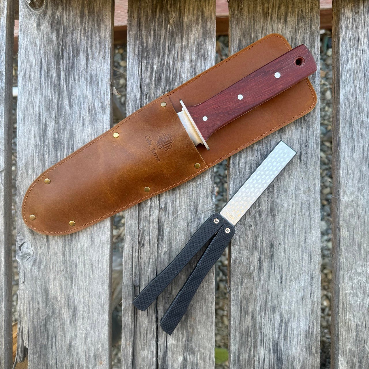 The Celtic Farm Hori Hori Garden Gift Box features a fixed-blade garden knife with a wooden handle, its brown leather sheath, and a black-handled diamond sharpener, all displayed on a weathered wooden surface.