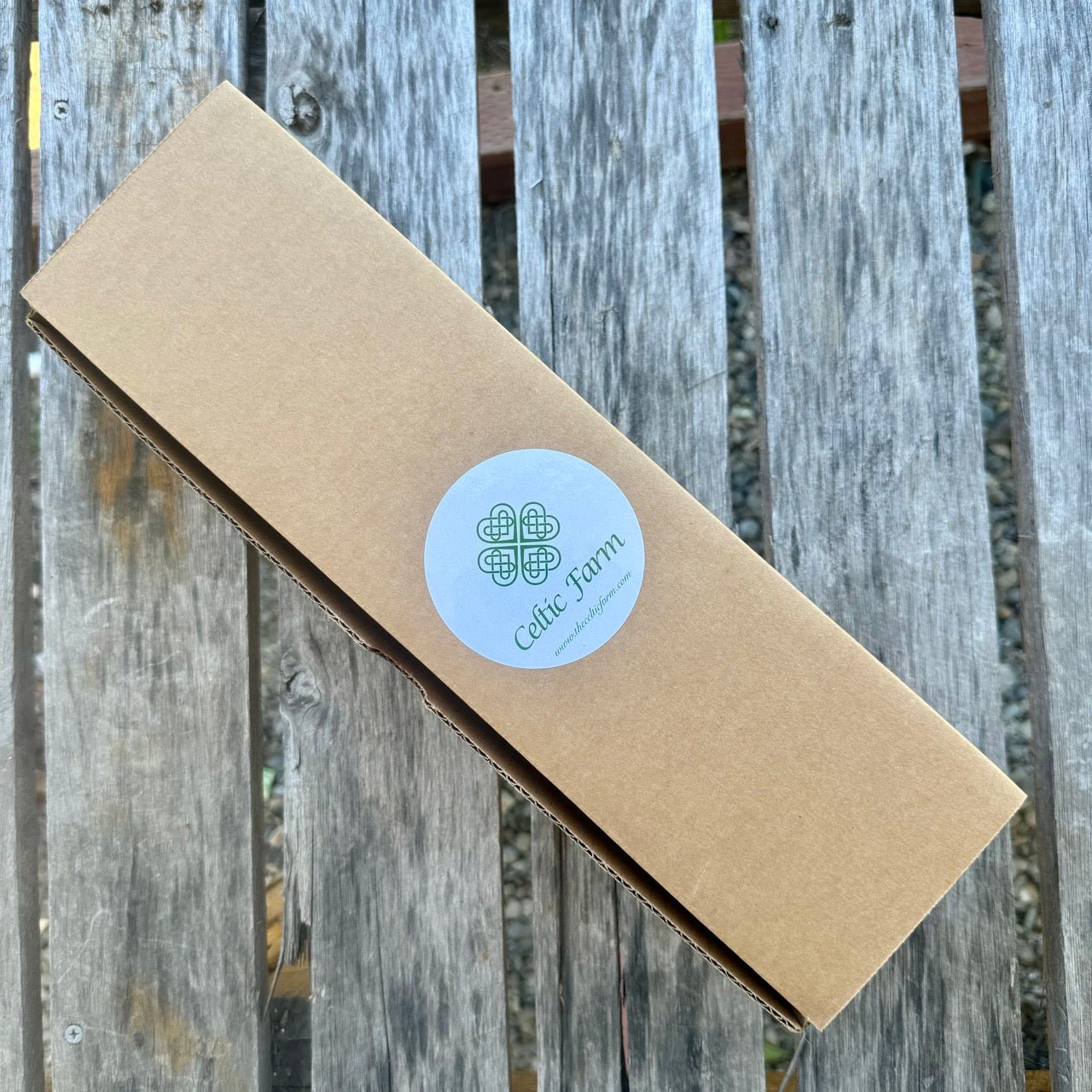 A long rectangular Hori Hori Garden Gift Box from The Celtic Farm, featuring a round logo, rests on a weathered wooden surface outdoors—ready to unveil the perfect garden knife for your next gardening adventure.