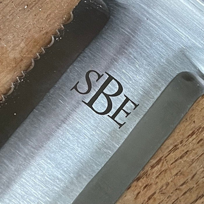Close-up of The Celtic Farms Our Katana Hori Hori Garden Tool blade engraved with SBE, showing its serrated edge and a wooden surface in the background.