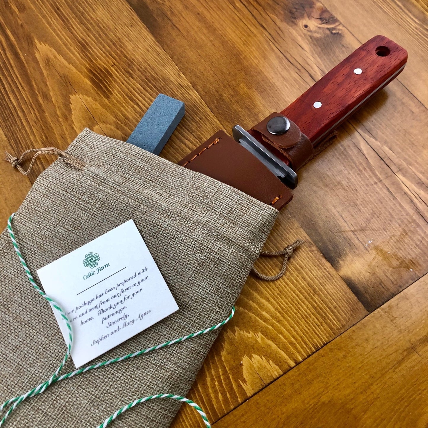 The Celtic Farms Our Katana Hori Hori Garden Tool, featuring a wooden handle and leather sheath, rests partly in a burlap pouch on wood. A blue sharpening stone and a card with text and a clover logo are beside this versatile gardening tool.