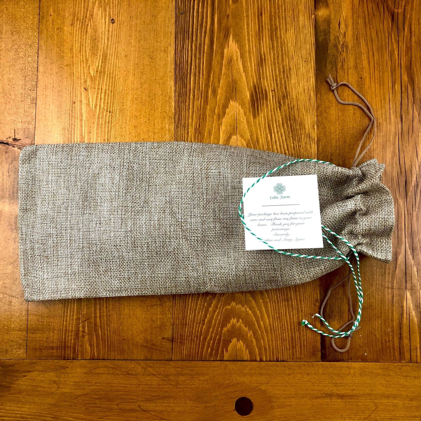 A burlap drawstring bag with a green and white tag, secured by a matching twisted string, sits on a wooden surface—ideal for storing The Celtic Farm’s Katana Hori Hori Garden Tool.
