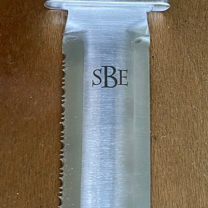 Close-up of The Celtic Farms Katana Hori Hori Garden Tool blade with serrated edge and sBE engraved, resting on a wooden surface.