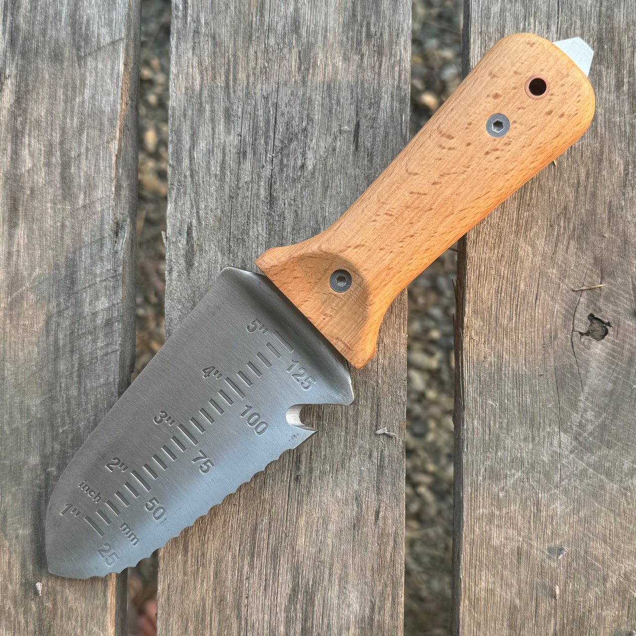 The Celtic Farms Our Tanto Hori Hori Garden Tool, with a wooden handle and stainless steel blade featuring measurement markings, serrated edge, and pointed tip, rests on a weathered wooden surface.