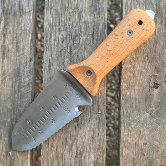 The Celtic Farms Our Tanto Hori Hori Garden Tool, with a wooden handle and stainless steel blade featuring measurement markings, serrated edge, and pointed tip, rests on a weathered wooden surface.