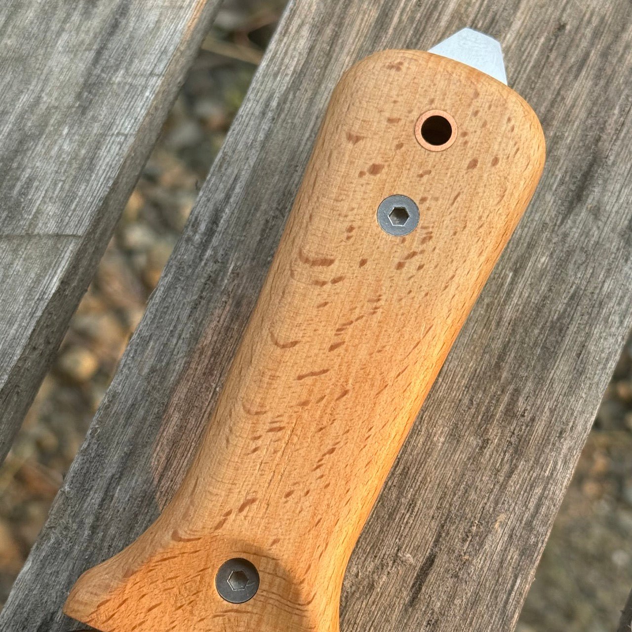 A close-up of The Celtic Farm’s Our Tanto Hori Hori Garden Tool handle, featuring two embedded metal hex nuts and a rounded silver cap, resting on a weathered wooden surface.