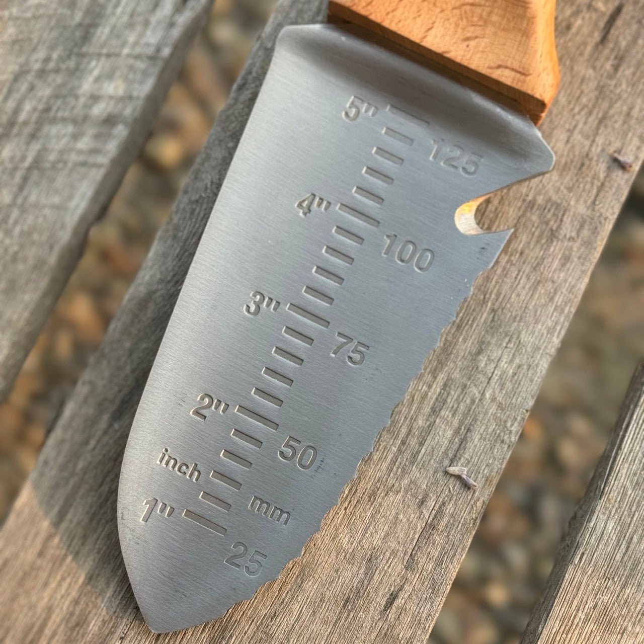 A close-up of The Celtic Farm’s Our Tanto Hori Hori Garden Tool, featuring a wooden handle and blade with inch and millimeter markings, displayed on a wooden surface.