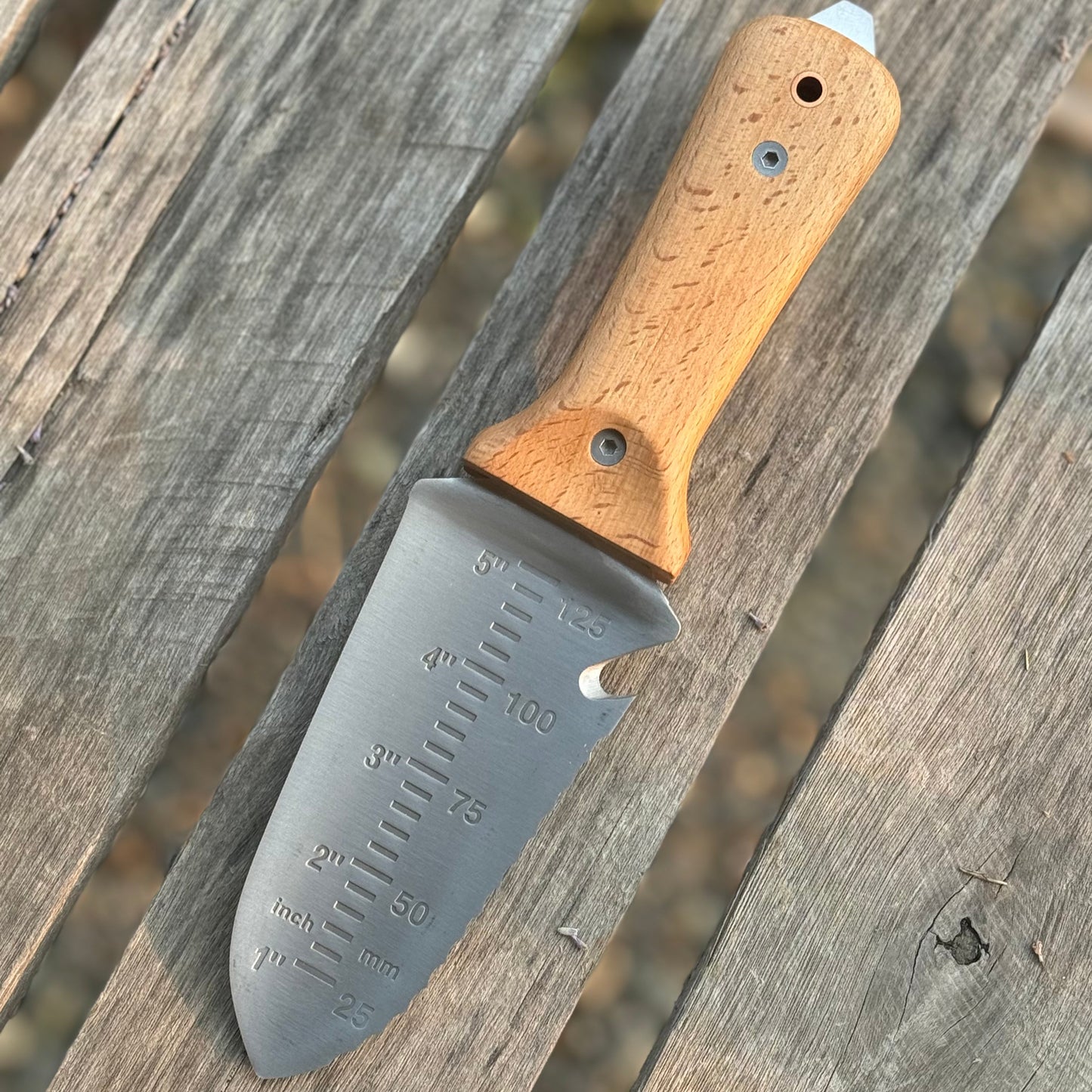 The Celtic Farm’s Tanto Hori Hori Garden Tool, featuring a wooden handle and stainless steel blade with inch and millimeter markings, is a must-have for any Japanese gardener, shown here on a weathered wooden surface.
