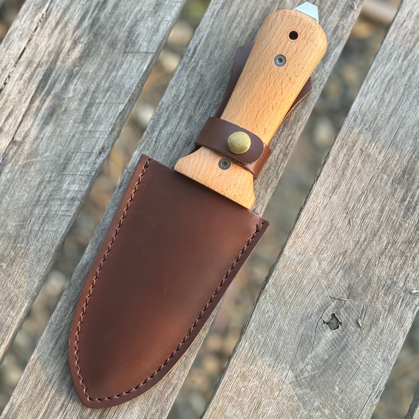 The Celtic Farm’s Our Tanto Hori Hori Garden Tool features a wooden handle, metal blade, and brown leather sheath, resting on a weathered wood surface—a traditional Japanese gardening essential ready for any task.