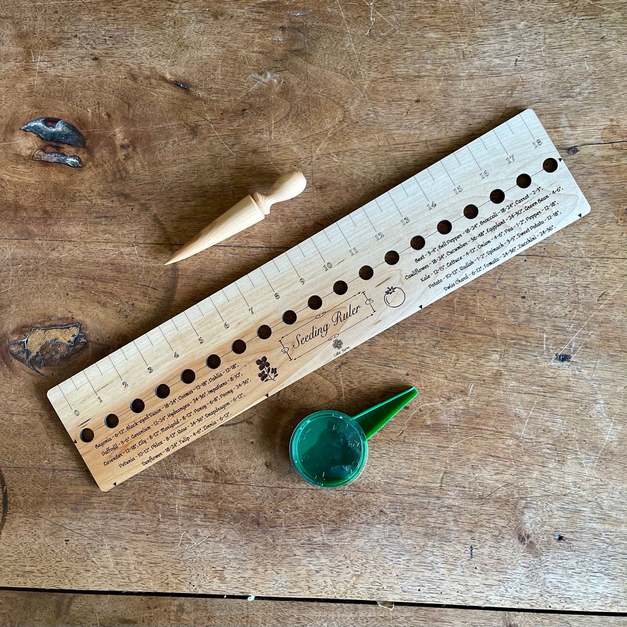 The Celtic Farm Seeding Gift Set - Dibble, Seeder, and Ruler includes a wooden ruler with measurements and planting holes, a wooden dibber, and a small green container—perfect gardening tools arranged on a wooden surface.