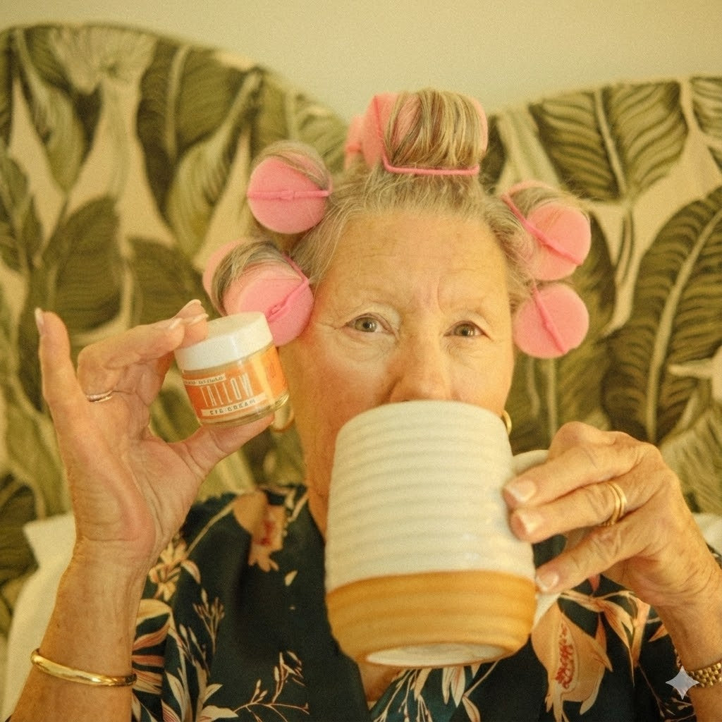 An older woman with pink hair rollers holds a mug and Lady May Tallow’s Natural Tallow Eye Cream | Caffeine Under Eye Cream, sitting in her robe before a leafy backdrop, ready to target under-eye puffiness.