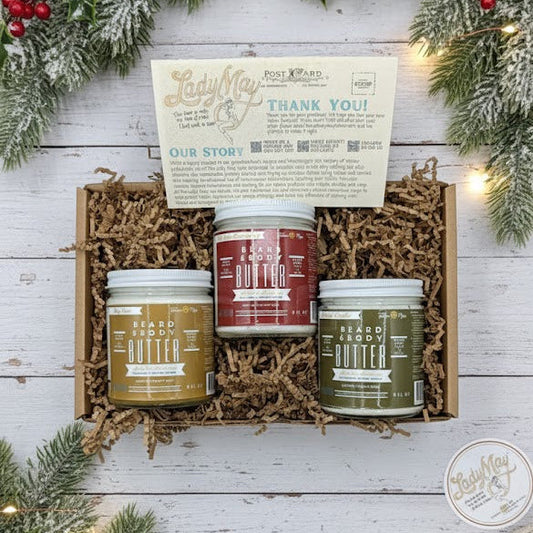 The Tallow Man Men’s Beard Butter Trio Gift Box by Lady May Tallow features three jars of 100% natural tallow beard butter, accented with festive greenery, berries, shredded paper, and a thank-you card on a white wood background.