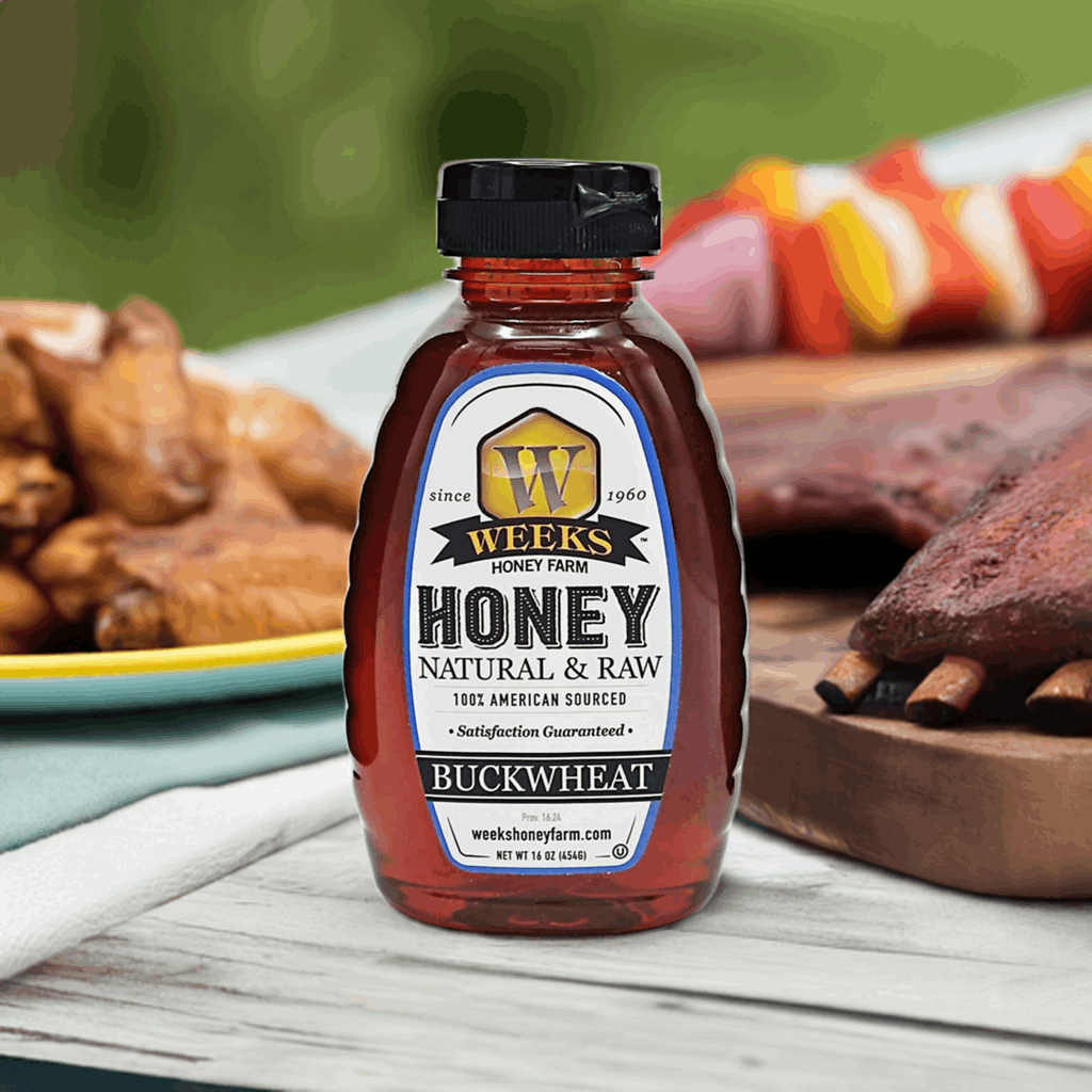 A bottle of Weeks Raw Buckwheat Honey by Weeks Honey Farm, pure and antioxidant-rich with a dark molasses flavor, sits on an outdoor table, while grilled ribs, chicken wings, and veggie skewers blur in the background.