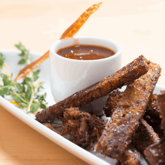 On a white rectangular plate, crispy breaded strips are served with herbs and a cup of dark dipping sauce made with Weeks Raw Buckwheat Honey from Weeks Honey Farm, topped with a shard of caramelized sugar.