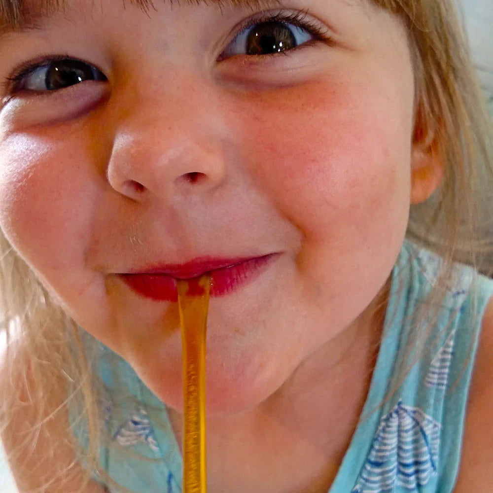 A young child with light brown hair and big eyes smiles while sipping from a yellow Weeks Honey Farm Raw Honey Straw (Orange Blossom, Wildflower, or Raspberry Honey), wearing a light blue sleeveless top.
