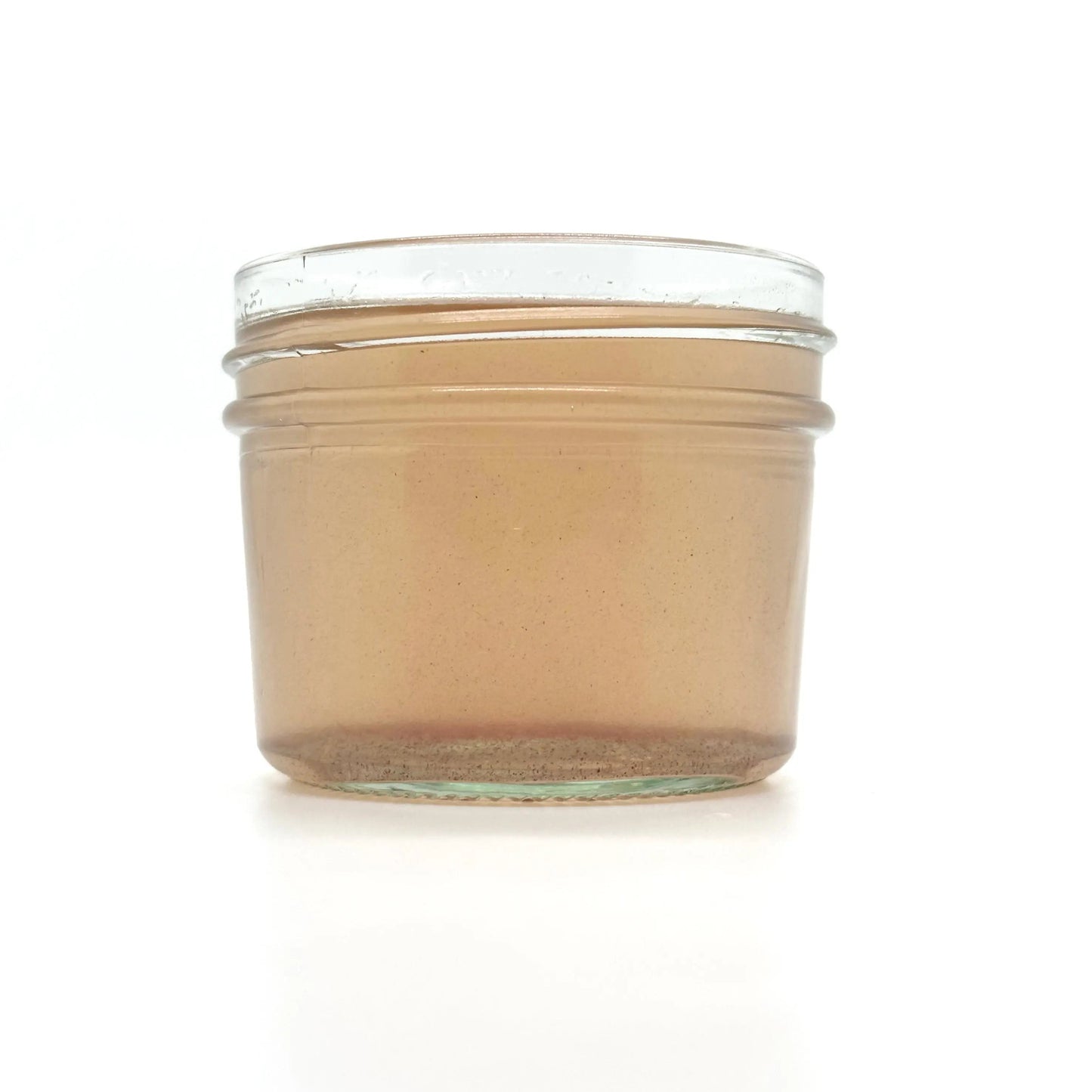 A small, clear glass jar of Rose Coconut Oil by Your Only Earth rests against a plain white backdrop, revealing its light brown, slightly translucent contents with hints of roses and visible specks.