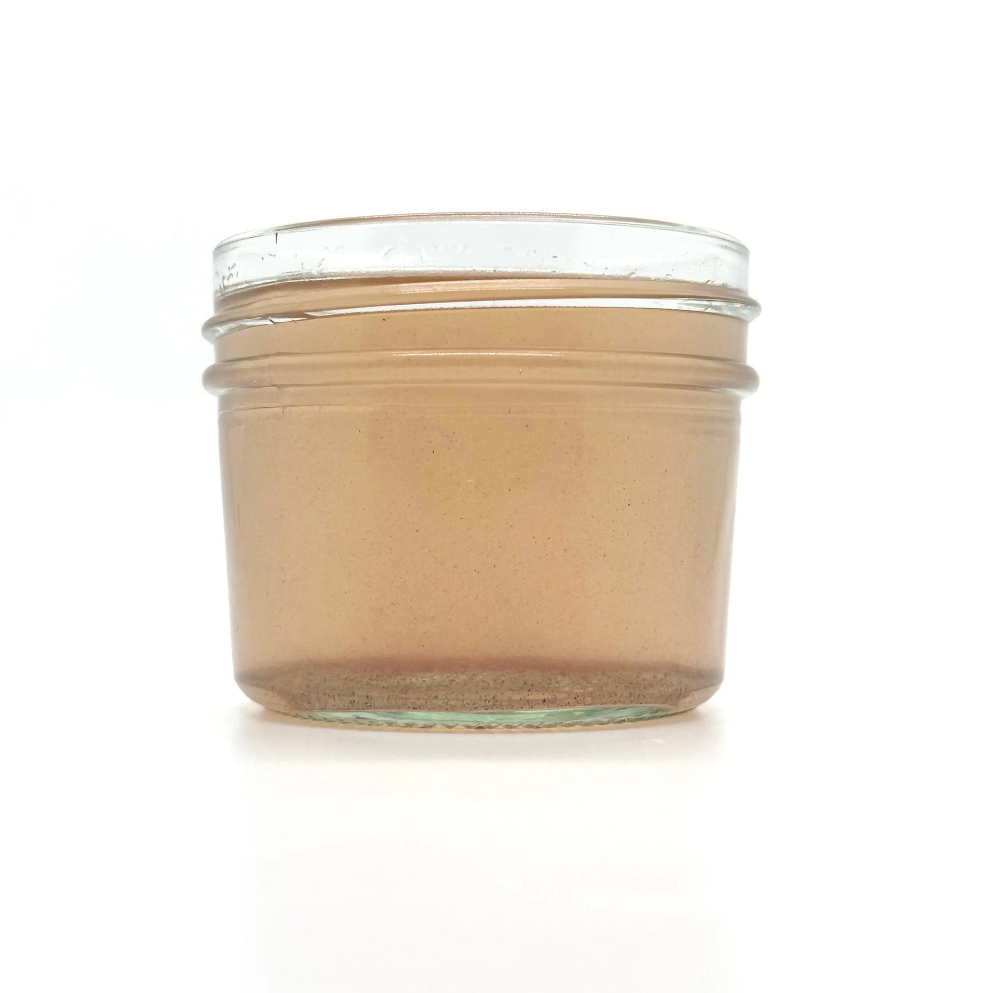 A small, clear glass jar of Rose Coconut Oil by Your Only Earth rests against a plain white backdrop, revealing its light brown, slightly translucent contents with hints of roses and visible specks.