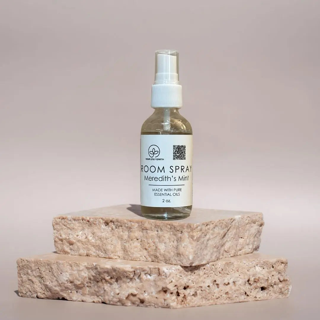 A transparent bottle labeled Room Spray: Your Only Earth with pure essential oils rests on stacked stone slabs against a plain background, featuring a white nozzle that enhances its elegant simplicity—an essential addition to your room spray collection.