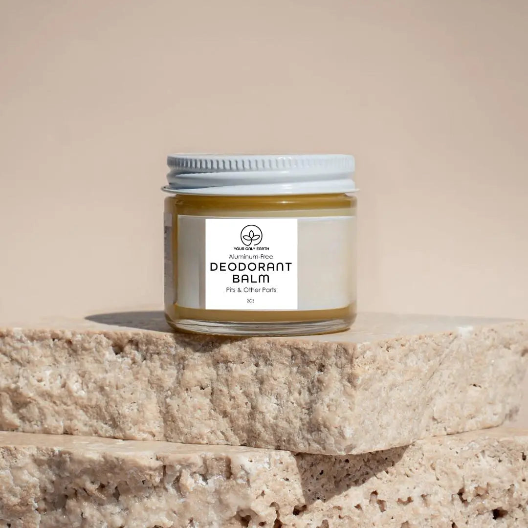 A glass jar of Your Only Earths Deodorant Balm with a white lid sits on beige stone slabs. The label reads Aluminum-Free Deodorant Balm with Essential Oils, Pits & Other Parts, 2oz, while the blurred background accentuates its natural and gentle vibe.