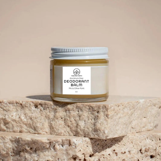 A glass jar of Your Only Earths Deodorant Balm with a white lid sits on beige stone slabs. The label reads Aluminum-Free Deodorant Balm with Essential Oils, Pits & Other Parts, 2oz, while the blurred background accentuates its natural and gentle vibe.