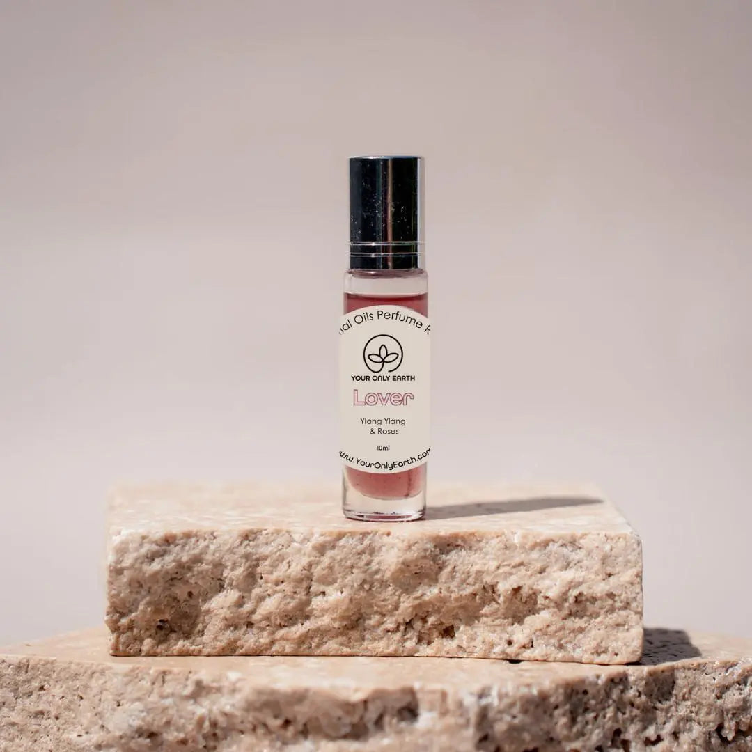 On two beige stone slabs stands a small, clear glass Perfume Roll-on by Your Only Earth with a black cap. The label Lover highlights an enchanting blend of ylang ylang and rose essential oils. The background is a neutral, soft focus.