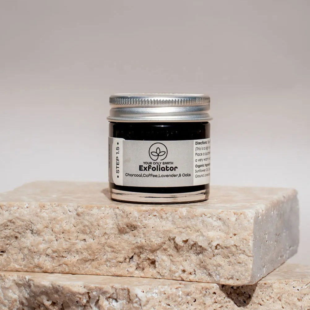 A small jar of Your Only Earth Exfoliator with a silver lid rests on beige stone tiles. The label features activated charcoal, coffee, lavender, and oats—ingredients prized for their exfoliating cleanser benefits.