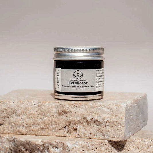 A small jar of Your Only Earth Exfoliator with a silver lid rests on beige stone tiles. The label features activated charcoal, coffee, lavender, and oats—ingredients prized for their exfoliating cleanser benefits.