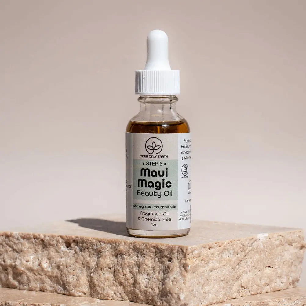A glass dropper bottle of Maui Magic Beauty Oil by Your Only Earth, featuring a golden liquid and white cap, stands on textured stone with a neutral, softly lit background. Ideal for soothing acne and rosacea.