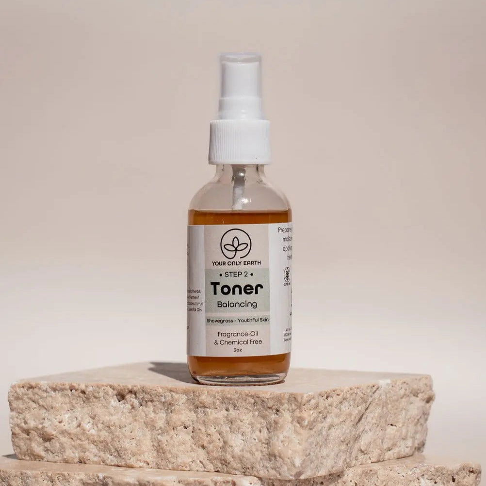 A clear spray bottle labeled "Toner" from Your Only Earth stands upright on stacked beige stone slabs against a light beige background. The label states it’s a botanical toner that’s free from fragrance, oil, and chemicals.