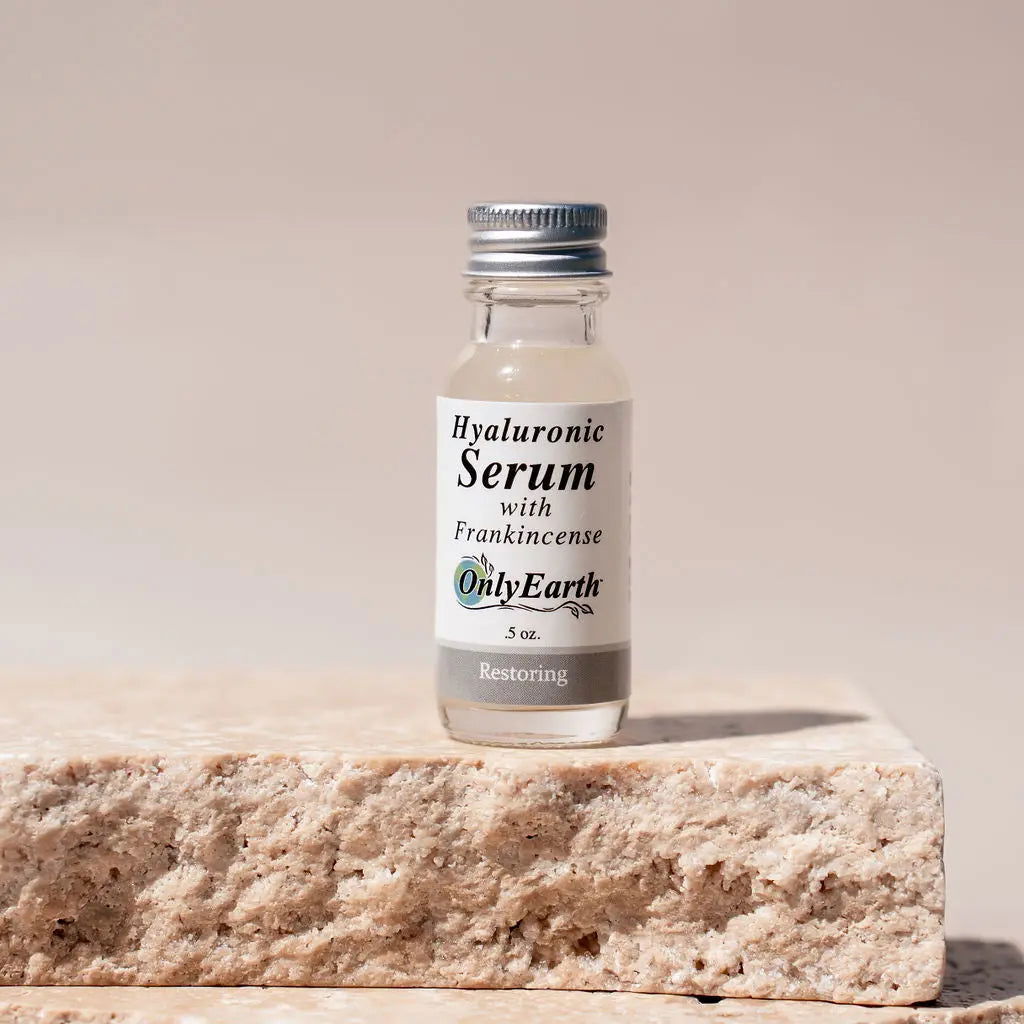 A small glass bottle labeled "Hyaluronic Acid Serum" by Your Only Earth, with a silver cap and white label, rests on a textured stone surface against a neutral background. This serum supports moisture retention for radiant skin.