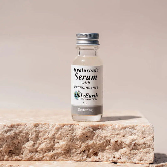 A small glass bottle labeled "Hyaluronic Acid Serum" by Your Only Earth, with a silver cap and white label, rests on a textured stone surface against a neutral background. This serum supports moisture retention for radiant skin.