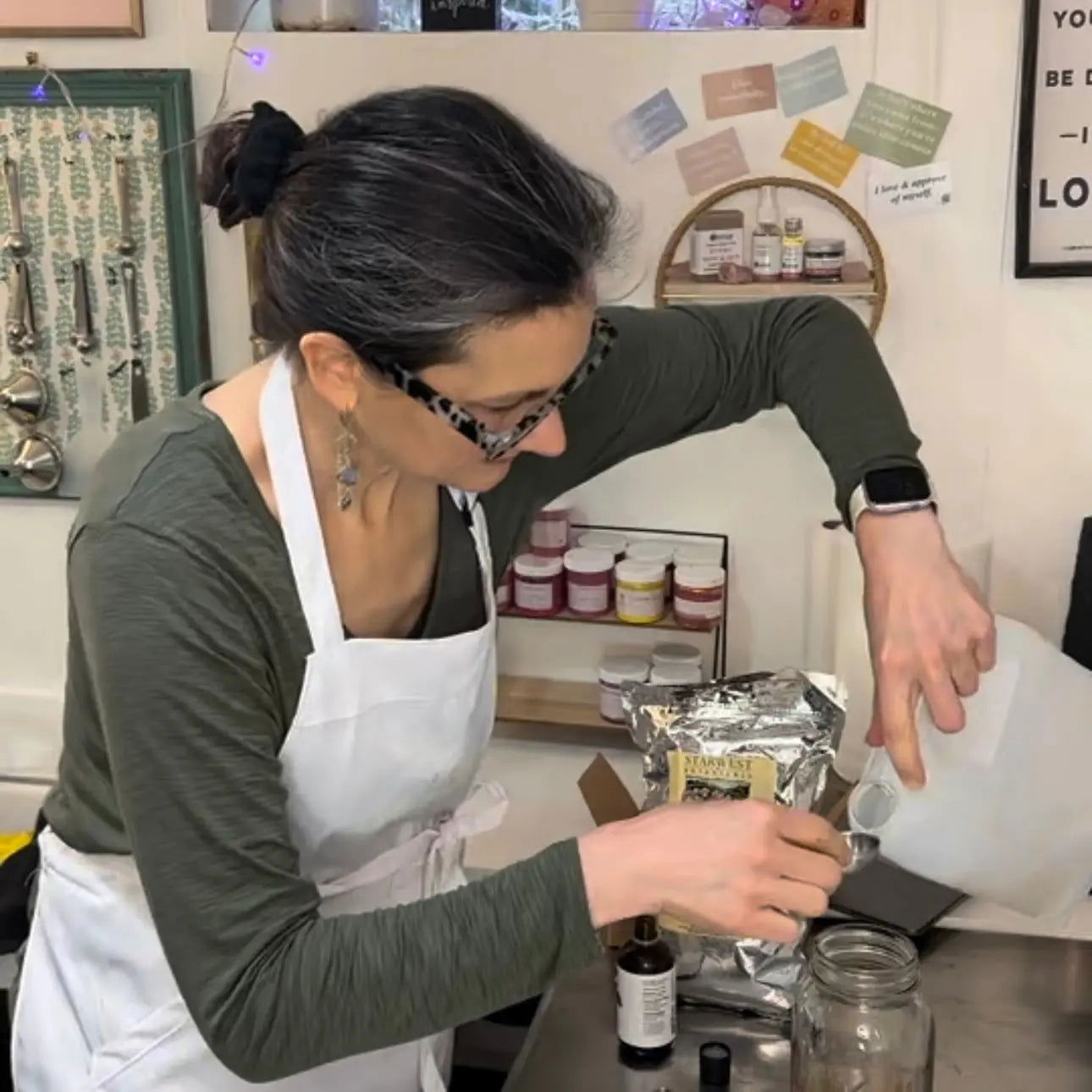 A person with a radiant complexion, glasses, earrings, and a white apron pours Your Only Earth Hyaluronic Acid Serum—known for moisture retention—into a glass jar in a kitchen or workshop filled with jars and utensils.