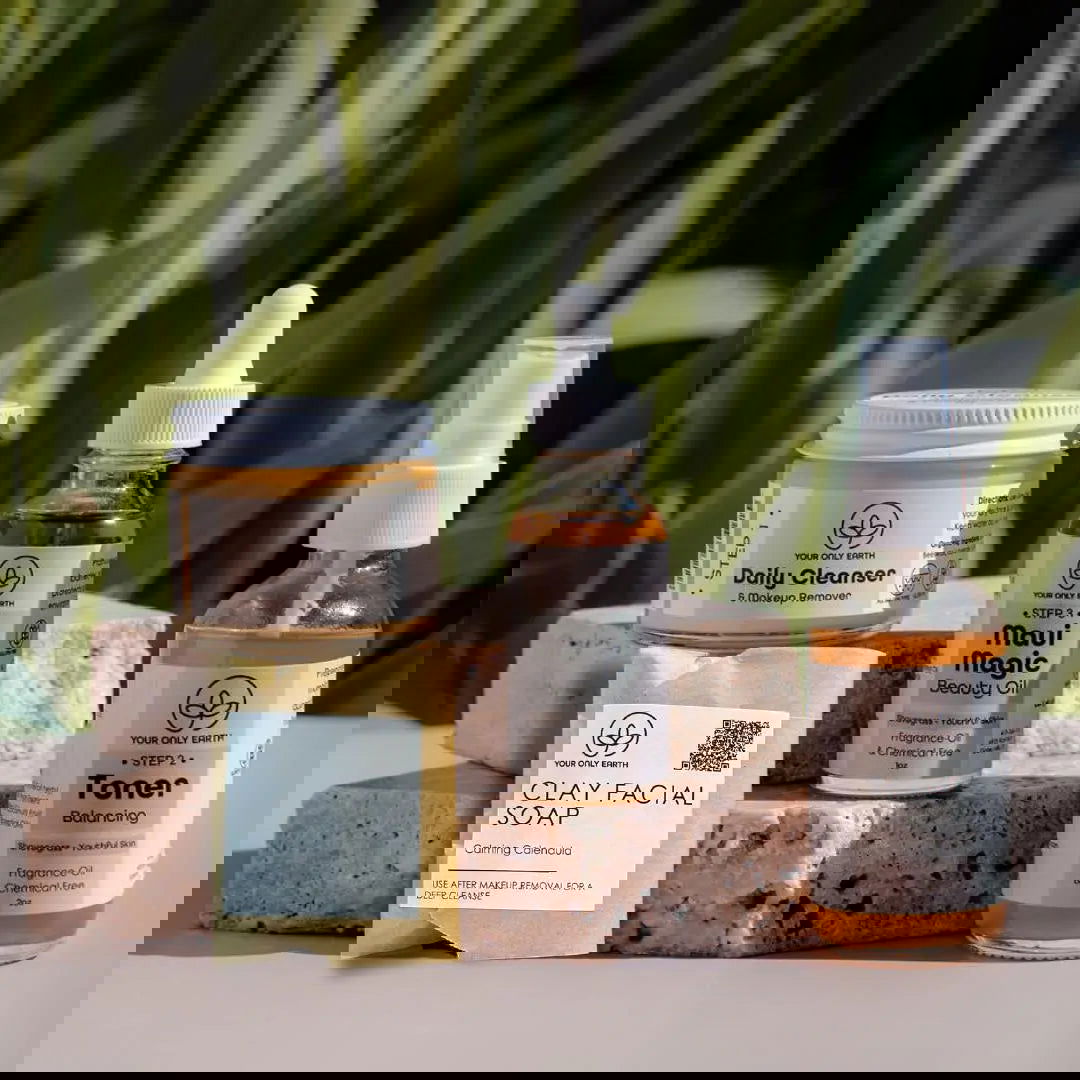 A Dewy Skincare Set by Your Only Earth, featuring artisan soap bars and jars with sustainable ingredients and decorative logos, is arranged on a white surface as green plant leaves blur in the foreground.