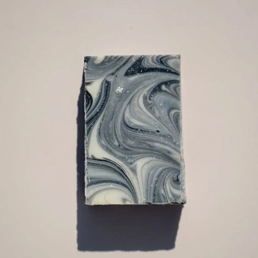 Farmer Toms Soap by Your Only Earth is a rectangular bar with an elegant marble swirl pattern, infused with charcoal and tea tree. Displaying shades of gray, white, and black, it rests on a light background with a soft shadow extending from it.