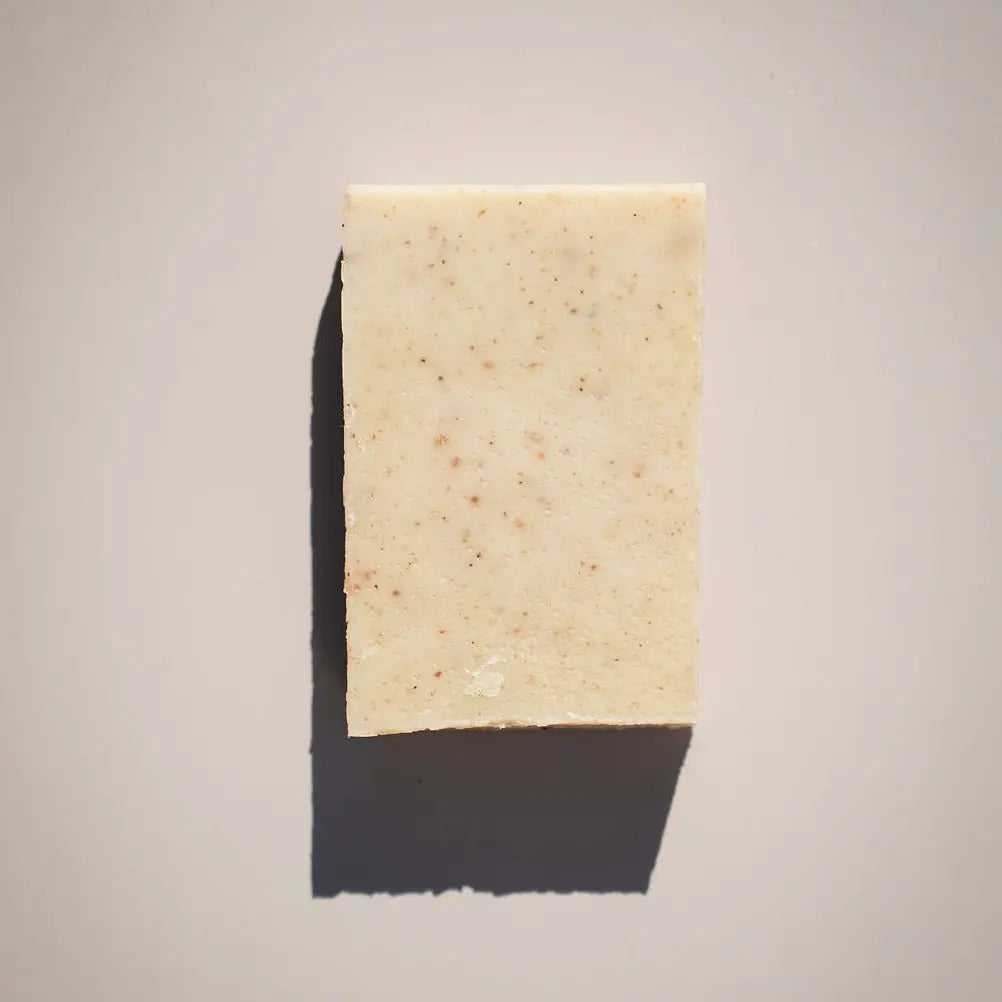 The Happy Dog Soap Bar by Your Only Earth, a rectangular speckled beige soap with oatmeal and organic oils, sits on a light gray surface. Its shadow is cast to the bottom left, revealing the lights direction.