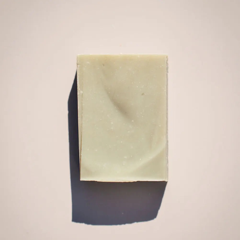 A minimalist image features Merediths Mint Soap by Your Only Earth, a light beige rectangular shaving bar with a smooth surface and tiny speckles suggesting French green clay, casting its shadow to the left on a plain background.